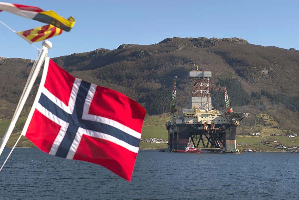 Norway national flag flying in front of a body of water with an oil rig and mountains in view.